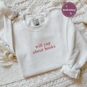 May include: White crewneck sweatshirt with the embroidered text "will yap about books" in red. The sweatshirt has long sleeves and a ribbed cuff. The image is on a white knitted surface.