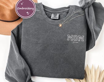 May include: A charcoal gray sweatshirt with the word "MOM" embroidered in white, with "Est. 2025" underneath. A gold necklace and a brown hat are also visible. The sweatshirt is made of a soft, comfortable material.