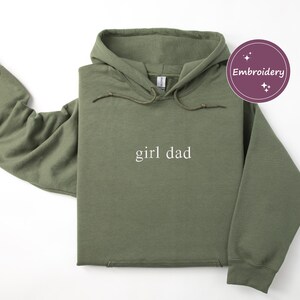 Puede incluir: Una sudadera con capucha verde oliva con las palabras "girl dad" en blanco, centradas en la parte delantera. La sudadera con capucha tiene una capucha con cord&oacute;n y mangas largas. Un gr&aacute;fico circular p&uacute;rpura con la palabra "Bordado" est&aacute; en la esquina superior derecha.