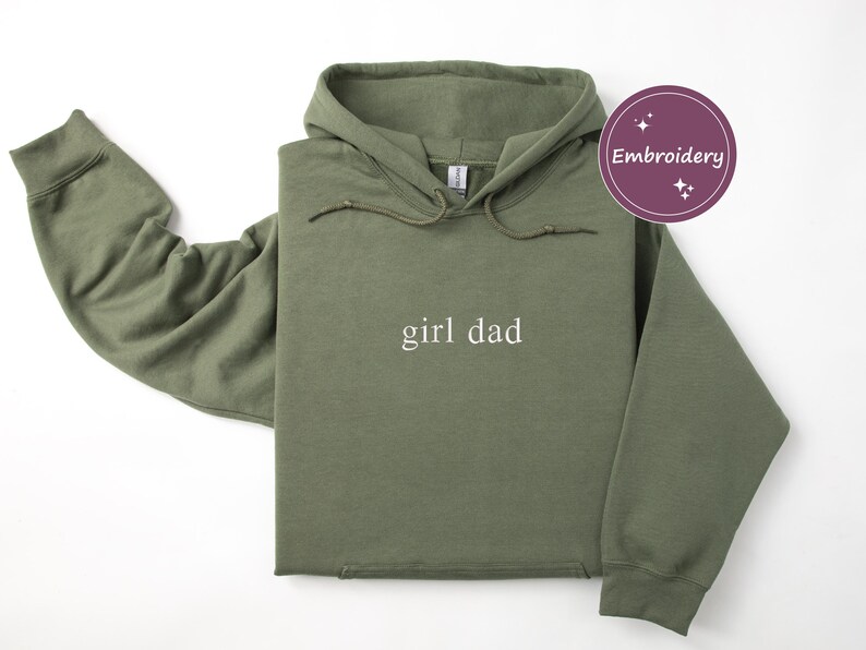 Puede incluir: Una sudadera con capucha verde oliva con las palabras "girl dad" en blanco, centradas en la parte delantera. La sudadera con capucha tiene una capucha con cord&oacute;n y mangas largas. Un gr&aacute;fico circular p&uacute;rpura con la palabra "Bordado" est&aacute; en la esquina superior derecha.