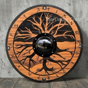 Op de afbeelding: Ronde houten schild met een zwart levensboomontwerp. Het schild heeft een zwarte metalen rand en een centrale baas. De tekst "The Decor Crest" is zichtbaar. Het schild is gemaakt van hout en metaal.