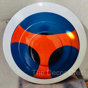 Puede incluir: Un objeto decorativo redondo con borde blanco, centro azul y diseño naranja. El centro es negro. El texto "The Decor Crest" está impreso en el objeto.