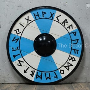 Peut inclure: Un bouclier rond en bois avec un bord noir, présentant des segments bleus et blancs alternés. Le bouclier a un bossage central noir et est orné d'inscriptions runiques noires sur le pourtour. Objet décoratif.