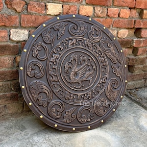 May include: A circular dark brown shield featuring a central relief carving of a two-headed dragon surrounded by intricate swirling patterns. The outer rim is accented with evenly spaced gold-coloured studs. The text The Decor Crest is visible.