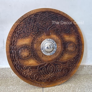 May include: A circular wooden shield with detailed carvings. The design includes a central tree motif, Norse knotwork, and runic inscriptions. A silver metal boss is in the centre. The Decor Crest is printed on the shield.