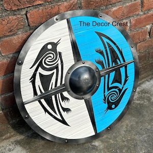 Peut inclure: Bouclier rond en bois avec une bordure et un bossage central en métal. Le bouclier est divisé en sections blanches et bleues, chacune avec un motif d'oiseau noir stylisé. Le texte "The Decor Crest" est visible sur la section bleue.