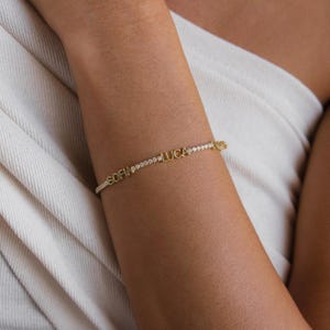 Op de afbeelding: Een goudkleurige armband met de namen "SOFIA" en "LUCA" in afzonderlijke letters. De armband is versierd met kleine, sprankelende steentjes. De armband bevindt zich op de arm van een persoon.