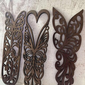 May include: Three intricately cut wooden butterfly decorations in varying designs. The butterflies are stained a warm brown color and feature detailed patterns. The central butterfly has a heart-shaped body. The background is a white, textured surface.