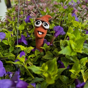 以下が含まれることがあります： 大きな目と尖った帽子を持つ、テラコッタ色の気まぐれなワームの彫刻。鮮やかな緑の葉と紫の花の中にあります。ワームは笑顔の表情をしています。彫刻は庭や植物のディスプレイに遊び心のあるタッチを加えます。