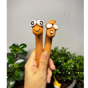 以下が含まれることがあります： 漫画の顔をした、オレンジ色のミミズ型の陶器製フィギュアが2つ。1つは笑顔、もう1つは驚いた表情をしています。フィギュアは手で持たれており、背景には緑の植物があります。庭の装飾品です。