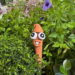 Könnte beinhalten: Ein skurriler, orangefarbener Keramik-Gartenstecker in Form eines Wurms mit großen Kulleraugen, einem kleinen Hut und einem lächelnden Mund. Der Stecker ist von üppigem, grünem Laub und lila Blüten umgeben.