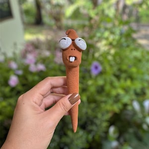 以下が含まれることがあります： 大きな白い縁取りの目と小さな開いた口を持つ、奇抜なテラコッタ色の生き物の彫刻。彫刻は丸い頭と先細りの体を持っています。背景はぼやけており、緑の葉と紫の花のヒントがあります。