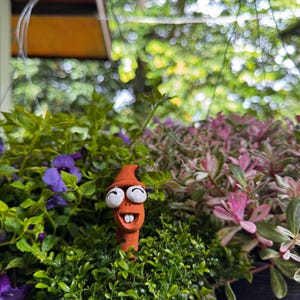 以下が含まれることがあります： 笑顔と大きな白い目を持つ、気まぐれなオレンジ色のセラミックガーデンステーク。ステークは、緑豊かな葉、紫の花、ピンクがかった葉のベッドに植えられており、活気のある庭のシーンを作り出しています。