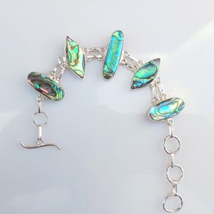 Op de afbeelding: Een zilveren armband met iriserende abalone schelpstukken in groen, blauw en paars. De armband heeft een tuimelsluiting en is verbonden door zilveren schakels. De abalone stukken zijn ovaal en langwerpig van vorm.