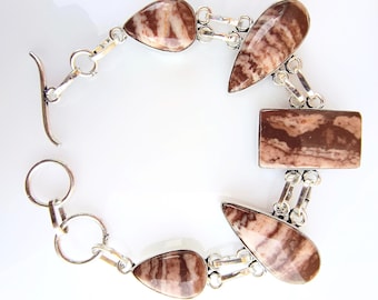 Op de afbeelding: Een zilveren armband met gepolijste, druppel- en rechthoekige stenen in bruin- en crèmetinten. De stenen zijn verbonden door zilveren schakels en een tuimelsluiting. De armband is ongeveer 18 cm lang.