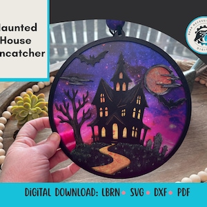 May include: A round, black haunted house suncatcher with a spooky scene. The image features a silhouette of a haunted house, bats, and a full moon against a purple and pink galaxy background. The text "Haunted House Suncatcher" is in the upper left.