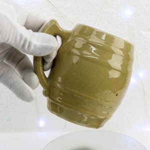 Puede incluir: Taza de cerámica verde oliva hecha a mano con un asa distintiva. La taza tiene una superficie texturizada y un diseño ligeramente en forma de barril. La taza está sostenida por una mano enguantada, mostrando su forma única y tonos terrosos.