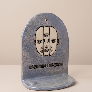 May include: A blue ceramic wall hanging with a shelf. The artwork features a graphic of three faces and the text "SELF-OBSERVATION IS SELF-CORRECTION" in black. The piece has a hole at the top for hanging.
