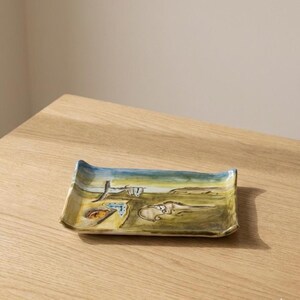 May include: Rectangular ceramic tray with a surrealist landscape design. The tray features a blue sky, a yellow and green landscape, and abstract shapes. The tray is placed on a wooden surface.