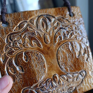 May include: A brown ceramic wall plaque with a detailed relief of a tree design. The tree has intertwined roots and branches, set against a textured background. A brown cord is attached for hanging. The plaque is square in shape.