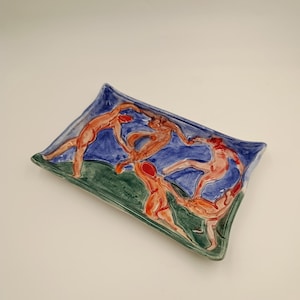 Op de afbeelding: Rechthoekige keramische schaal met een geschilderd ontwerp van figuren in tinten oranje en rood tegen een blauwe en groene achtergrond. De schaal heeft een licht verhoogde rand en een glanzende afwerking.