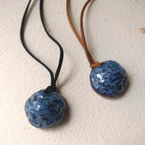 May include: Two necklaces featuring round, textured ceramic pendants with a glossy blue glaze and dark brown recessed details. One pendant hangs from a black cord, while the other hangs from a tan cord, both showcasing a unique, organic shape.