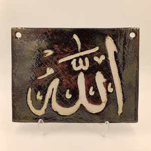Op de afbeelding: Rechthoekige keramische plaat met het Arabische woord "Allah" in witte kalligrafie tegen een donkere, gevlekte achtergrond. De plaat heeft twee gaten om op te hangen en wordt op een transparante standaard gepresent.