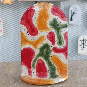 May include: A ceramic arch-shaped wall hanging with a colorful abstract design. The design features red, orange, and green shapes on a white background with black speckles. The bottom and top edges are orange.