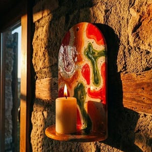 May include: A ceramic wall-mounted candle holder with a colorful abstract design in red, green, orange, and white. A lit, cream-colored candle sits on the holder's small shelf. The holder is mounted on a textured stone wall, illuminated by sunlight.