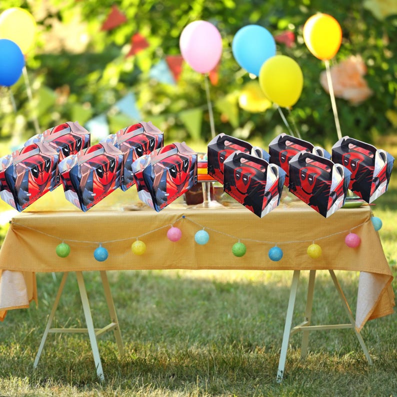 May include: A selection of Deadpool-themed party favour boxes with red and black graphics, arranged on a yellow tablecloth. The boxes have handles and are set up for a party, with balloons in the background.