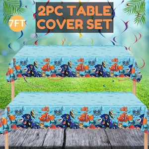 May include: Two-piece table cover set. The light blue table covers feature a repeating pattern of orange clownfish, sharks, and other sea creatures. The text "2PC TABLE COVER SET" is displayed in orange. The table covers are 7 feet long.