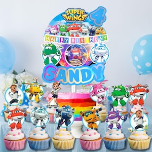 May include: A colorful birthday cake with a rainbow frosting, topped with a "Super Wings" cake topper and character cupcake toppers. The cake is decorated with the name "Sandy" and the number "4". Cupcakes with white frosting and character toppers are arranged around the cake.