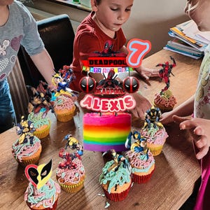 Puede incluir: Una escena de cumplea&ntilde;os con cupcakes y una tarta arco&iacute;ris. Los cupcakes est&aacute;n decorados con figuras de superh&eacute;roes y glaseado colorido. Un adorno para tarta dice "Deadpool Happy Birthday Alexis 7". La tarta arco&iacute;ris es el centro.