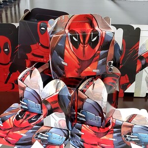 May include: A selection of Deadpool-themed party favour boxes. The boxes are predominantly red and black, showcasing the character's mask and costume design. The boxes are folded and assembled, ready for use.