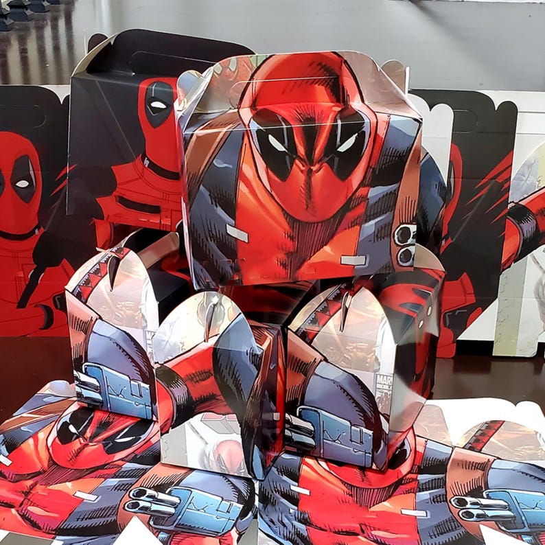 May include: A selection of Deadpool-themed party favour boxes. The boxes are predominantly red and black, showcasing the character's mask and costume design. The boxes are folded and assembled, ready for use.