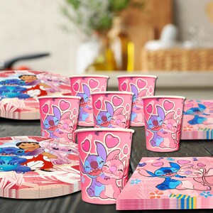 May include: A party supplies set featuring pink paper cups, plates, and napkins. The items are decorated with a cartoon character design in shades of pink, blue, and white, with heart accents. The set is arranged on a dark surface.