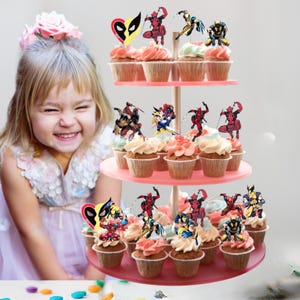 Puede incluir: Un soporte para cupcakes de tres niveles con cupcakes cubiertos con glaseado colorido y adornos tem&aacute;ticos de superh&eacute;roes. Los cupcakes est&aacute;n dispuestos en niveles rosas, con un ni&ntilde;o sonriente en el fondo.