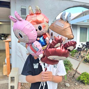 May include: A collection of foil balloons in various colors and character designs. The balloons include a pink bunny, a red-haired character in a blue outfit, and a brown-helmeted character. The balloons are held together with white ribbons.