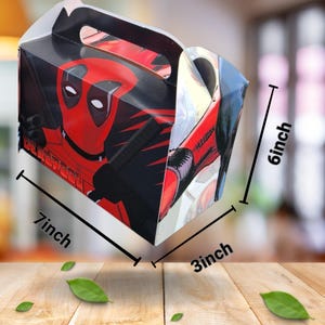 May include: A rectangular gift box featuring a Deadpool graphic. The box is red, black, and white, with dimensions of 17.78 x 7.62 x 15.24 cm. It has a handle for carrying.