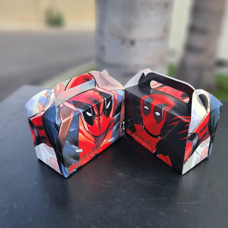 May include: Two gift boxes with a Deadpool comic book design. The boxes are predominantly red, black, and blue, with a handle. The boxes have a comic book style design and the word "DEADPOOL" is visible on one of the boxes.
