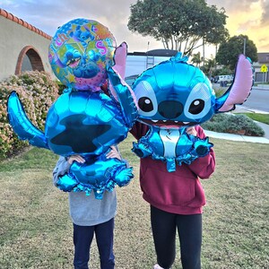May include: Two people holding large, blue foil balloons shaped like the character Stitch from the Disney movie Lilo & Stitch. One balloon is a round shape with Stitch's face and the words "Stitch" and "Aloha".
