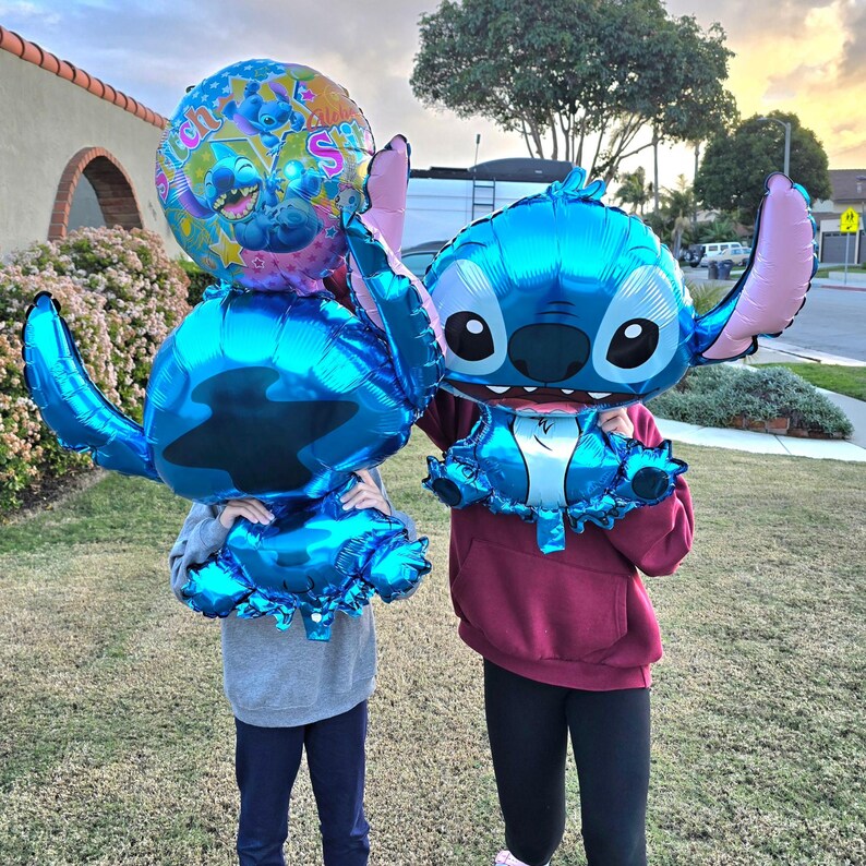 May include: Two people holding large, blue foil balloons shaped like the character Stitch from the Disney movie Lilo & Stitch. One balloon is a round shape with Stitch's face and the words "Stitch" and "Aloha".