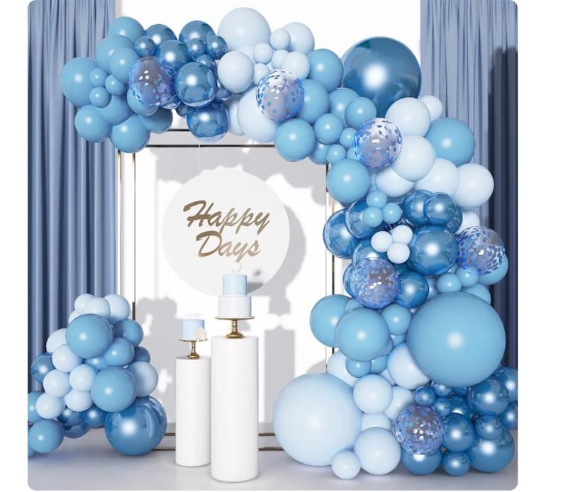 May include: A festive balloon arch in shades of blue and white, with a backdrop of blue curtains. The arch features various sizes of balloons, including some with a transparent, glittery effect. A sign with the words "Happy Days" is in the center.