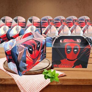 May include: A selection of Deadpool-themed gift boxes. The boxes are predominantly red and black, showcasing the Deadpool character's face. They are arranged on a wooden surface, accompanied by a red and white striped cloth.