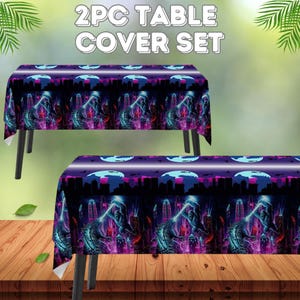 May include: A 2-piece table cover set featuring a vibrant design. The covers display a cityscape with a monster, in shades of purple, blue, and pink. The text "2PC TABLE COVER SET" is at the top. The table covers are on a wooden surface.