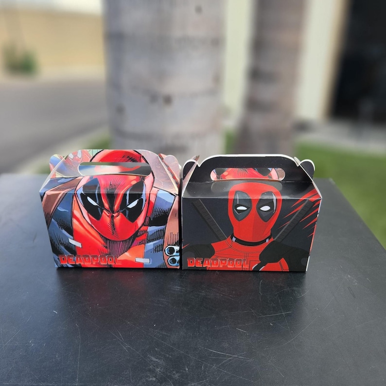 May include: Two Deadpool-themed gift boxes. One box features a comic-style Deadpool image with red, black, and blue colours. The other box has a black background with a red Deadpool image. Both boxes have the word "DEADPOOL" printed on them.