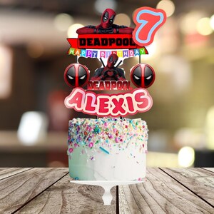 Puede incluir: Una tarta de cumplea&ntilde;os con glaseado blanco y chispas de colores, decorada con adornos tem&aacute;ticos de Deadpool. Los adornos incluyen una figura de Deadpool, el n&uacute;mero 7 y el nombre "Alexis". La tarta tambi&eacute;n tiene las palabras "Happy Birthday".