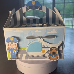 Puede incluir: Una caja de favor de helicóptero de policía azul y blanco con un policía y un perro policía. La caja tiene un asa a rayas blancas y negras y un sombrero de policía azul en la parte superior.