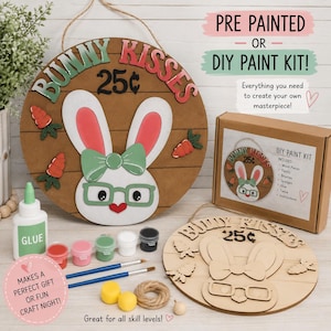 May include: A round wooden sign featuring a bunny face with green glasses and a bow, text reading BUNNY KISSES 25¢, and a DIY paint kit including a cardboard box, glue, paint pots, brushes, twine, and an unpainted wooden bunny sign for crafting.