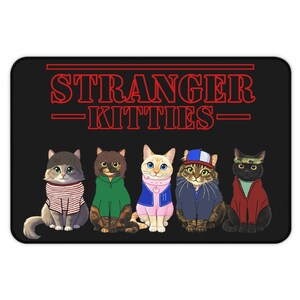May include: Black rectangular mat featuring five cartoon cats dressed in outfits. The mat has the text "STRANGER KITTIES" in red. The cats are wearing various outfits, including a striped shirt, a green hoodie, and a blue jacket.